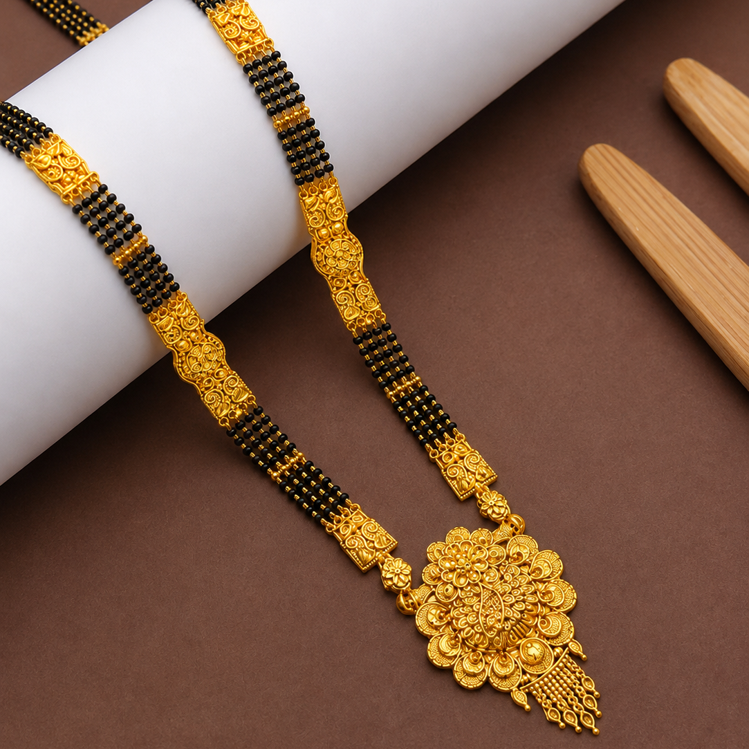 An ornate gold necklace with a detailed floral pendant and black-and-gold beaded chain, displayed on a white cylindrical prop over a brown background with wooden tools.